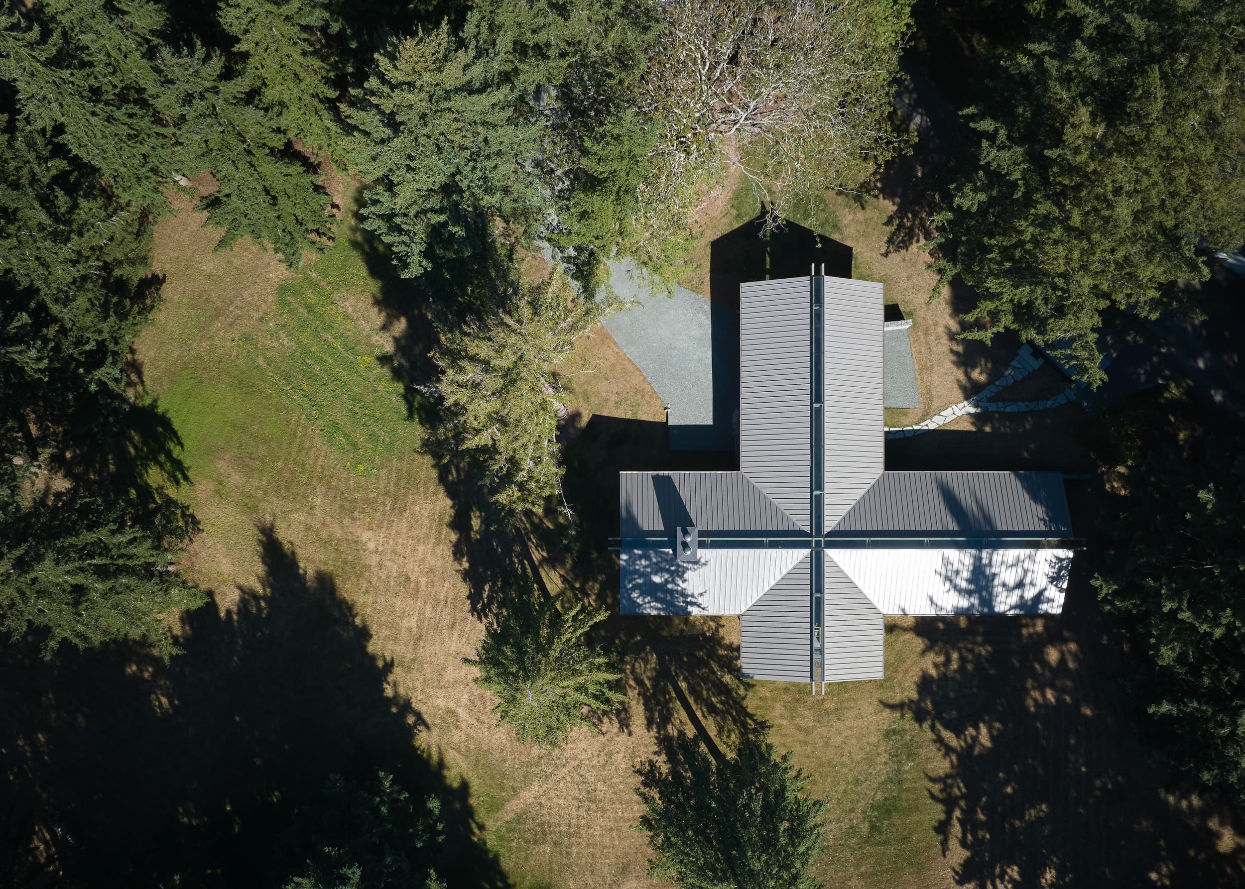 An aerial photograph of a modern house with a cross-shaped roof in a forest clearing.
