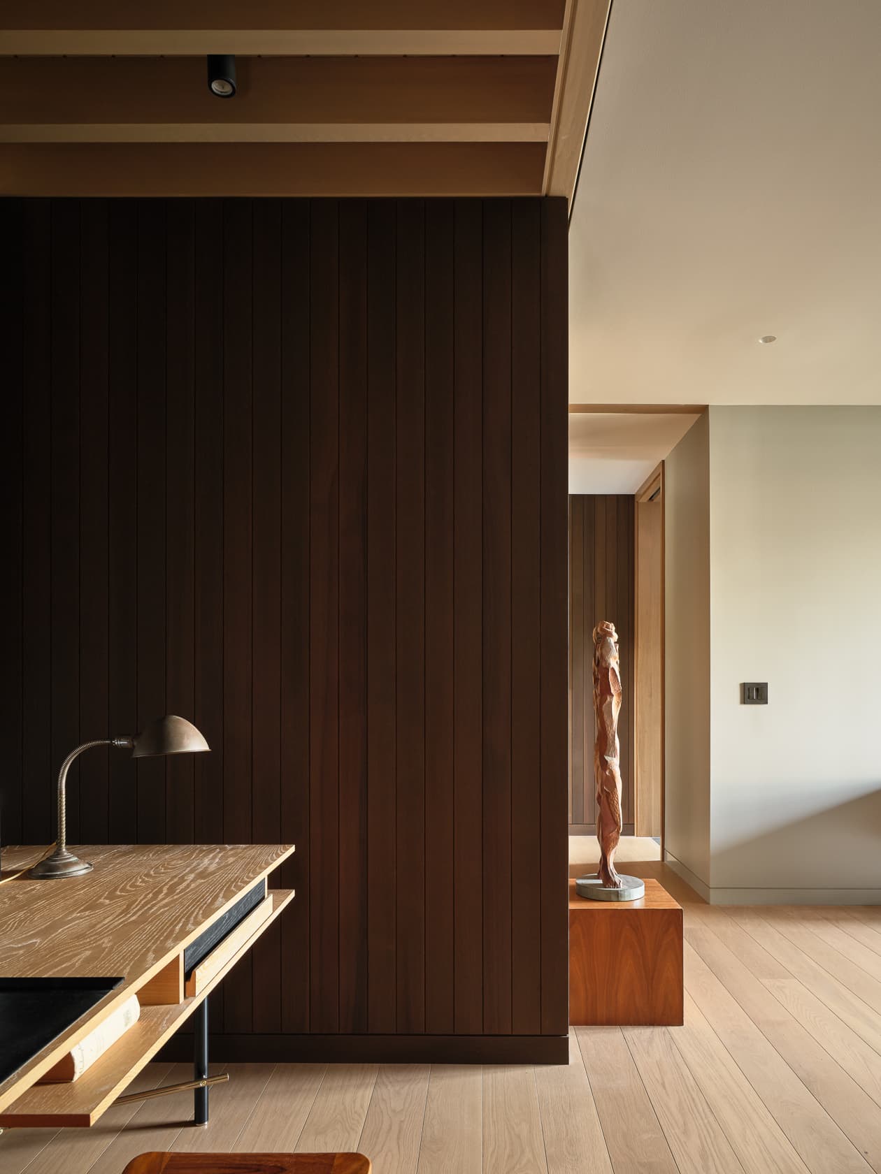 A modern room with a wooden desk, dark wood paneling, and a hallway with a sculpture.
