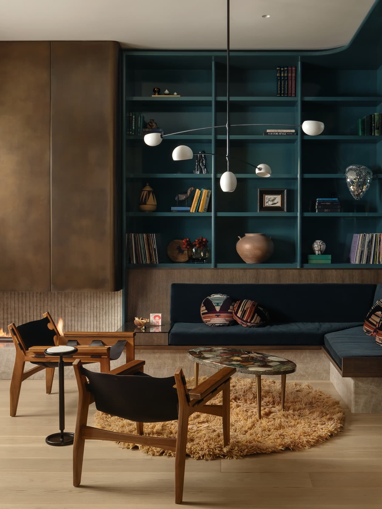 A modern lounge with a large teal bookshelf, a blue sofa, and wooden chairs on a shag rug.