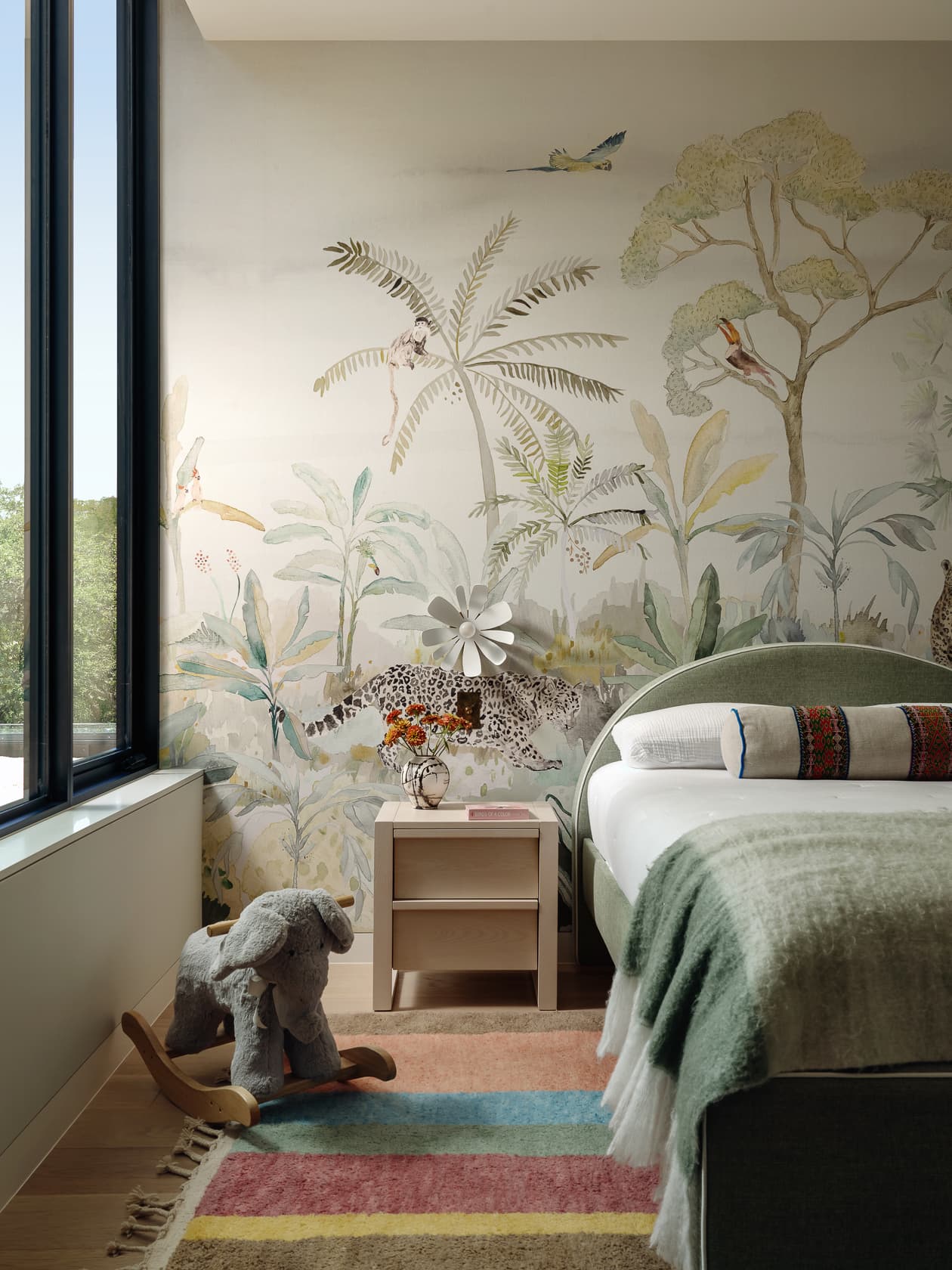A sunlit child's bedroom with a jungle-themed mural wallpaper and a plush rocking elephant.