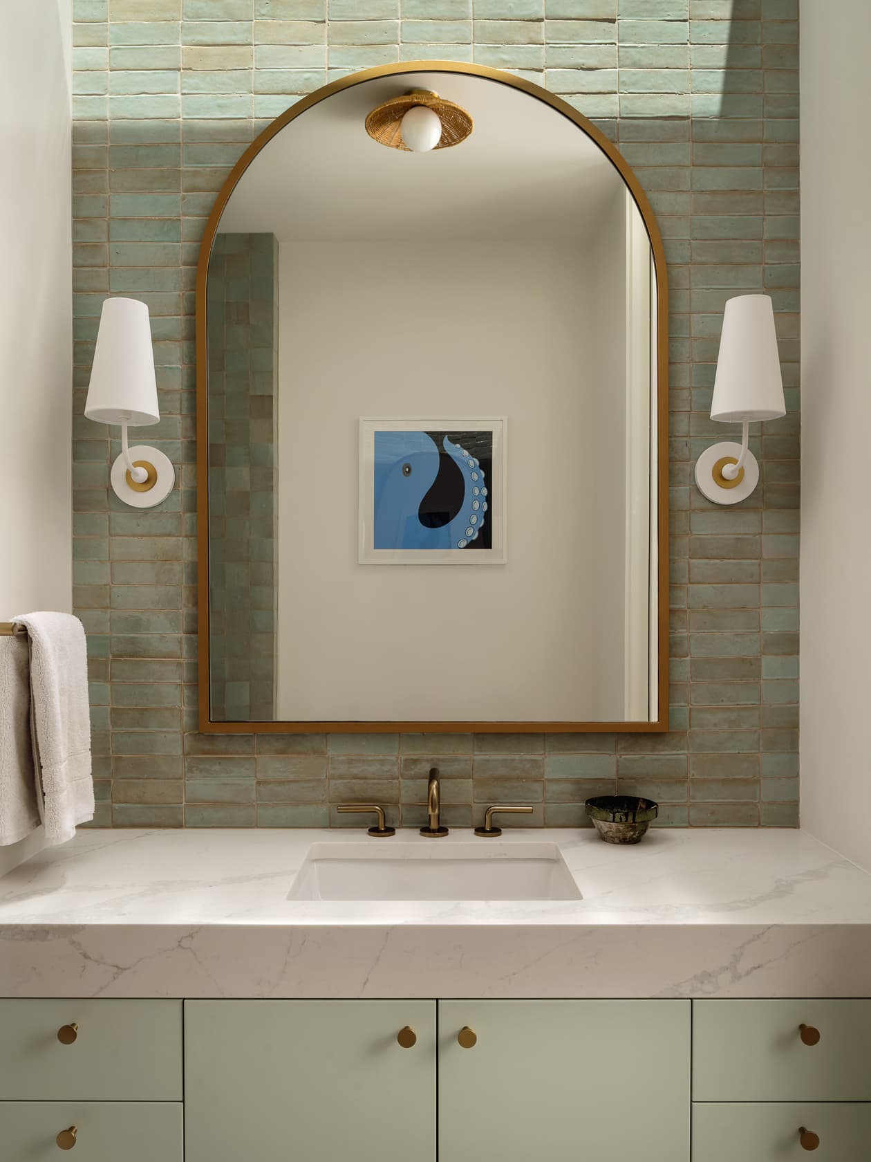 A gold arched mirror hangs over a sink with a green tile backsplash and sage green vanity.