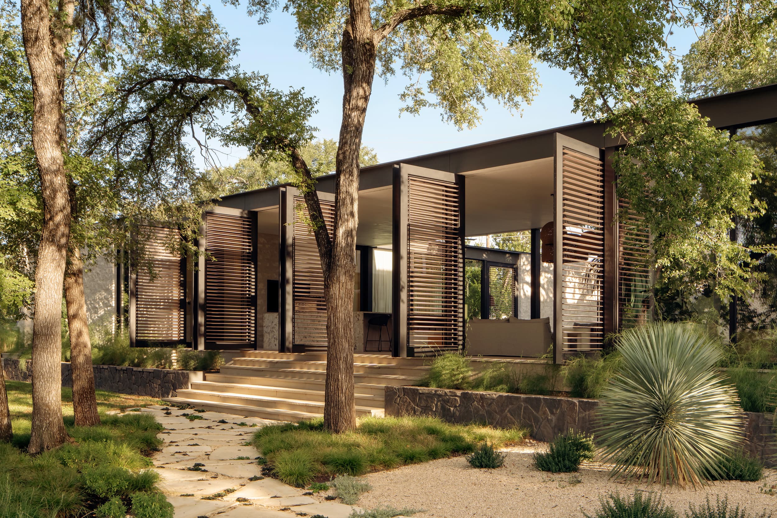 A modern house with large, slatted screen walls nestled in a wooded, landscaped yard.