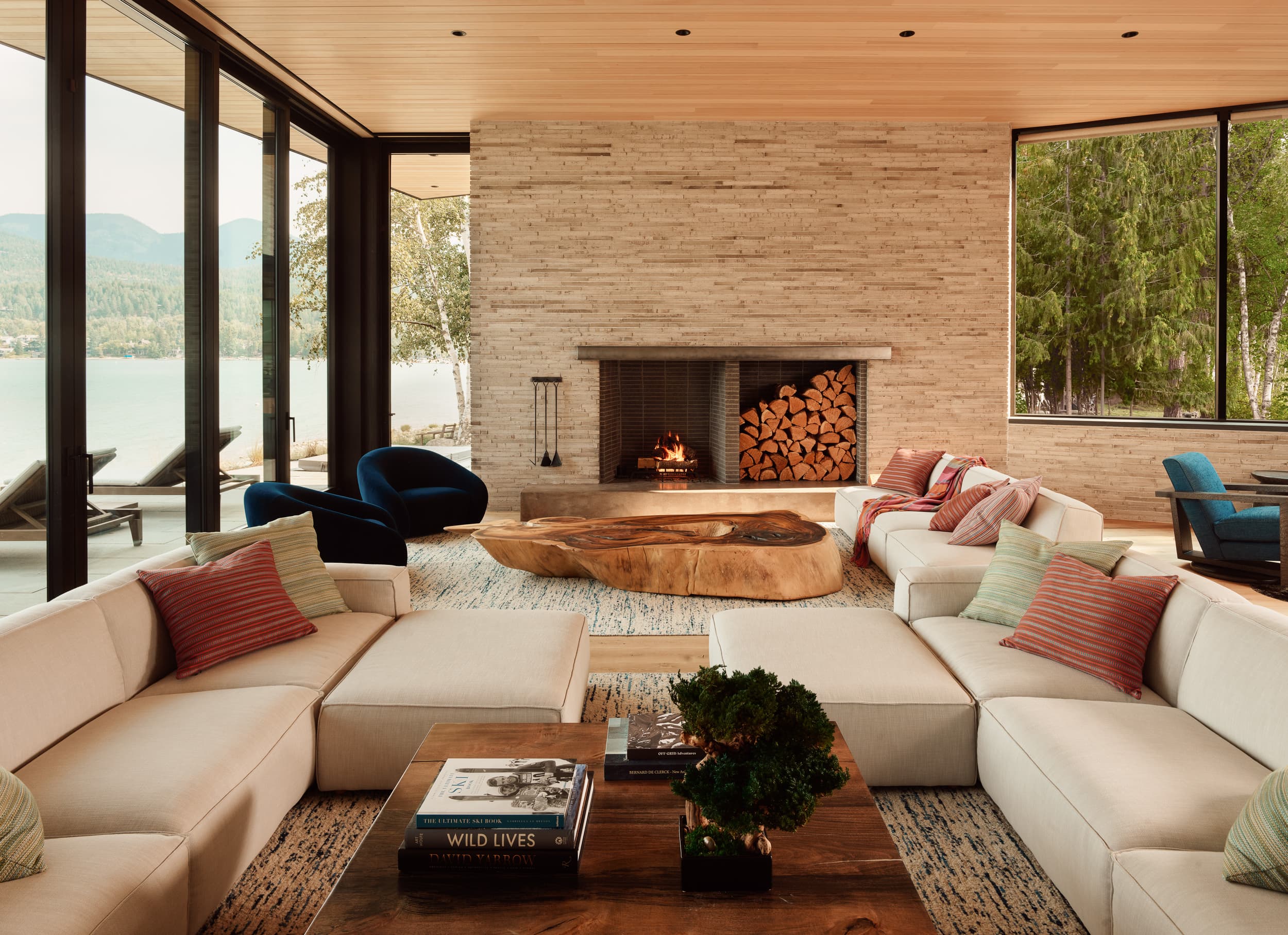 A modern living room with a large white sectional sofa, a stone fireplace, and windows overlooking a lake.