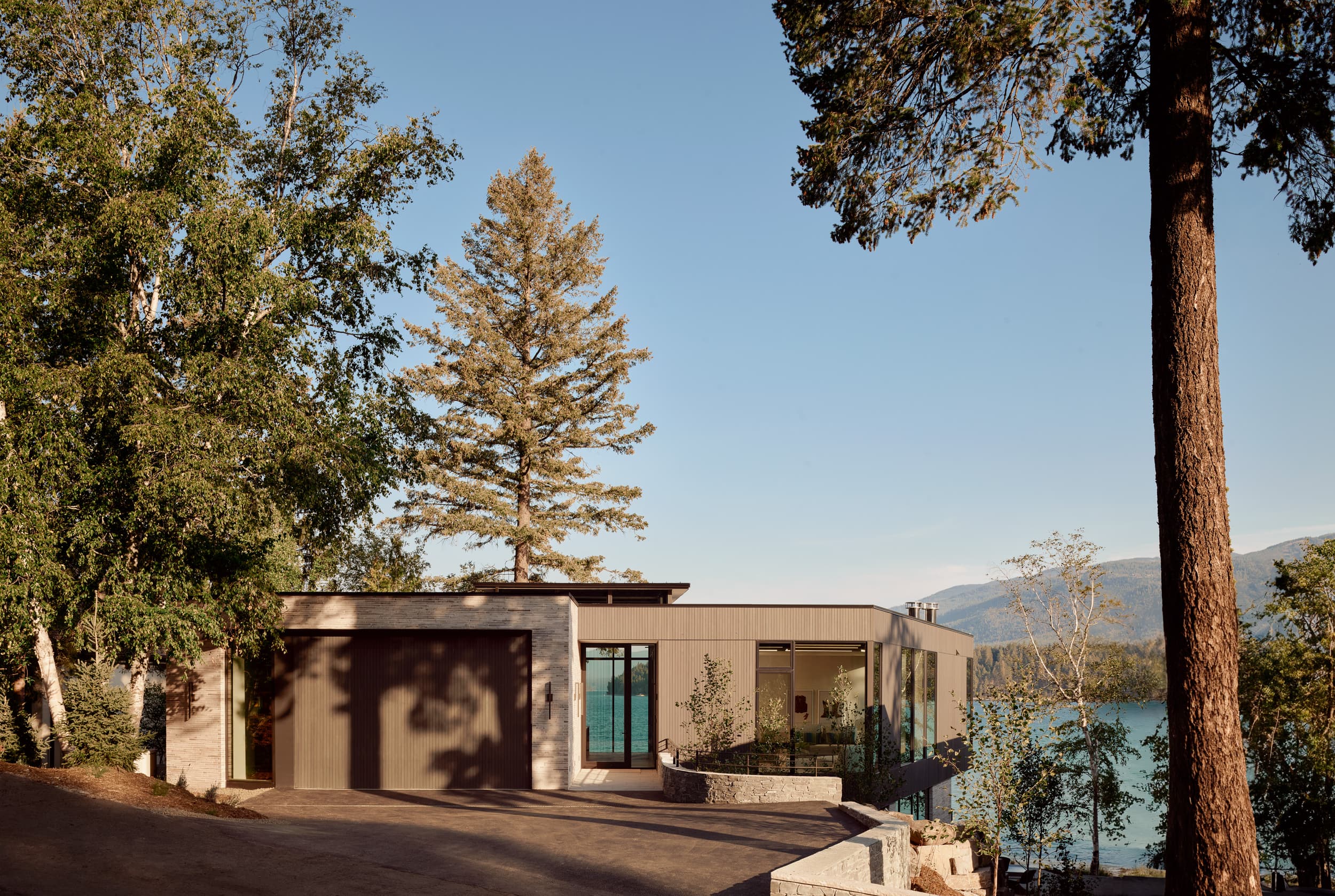 A modern, flat-roofed home with a garage, nestled in the woods beside a lake.
