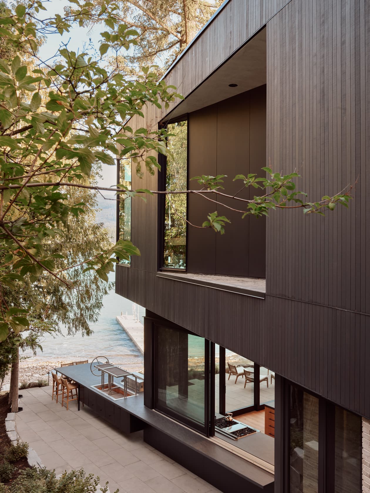 Exterior of a dark, modern house with a recessed balcony, overlooking a waterfront patio.