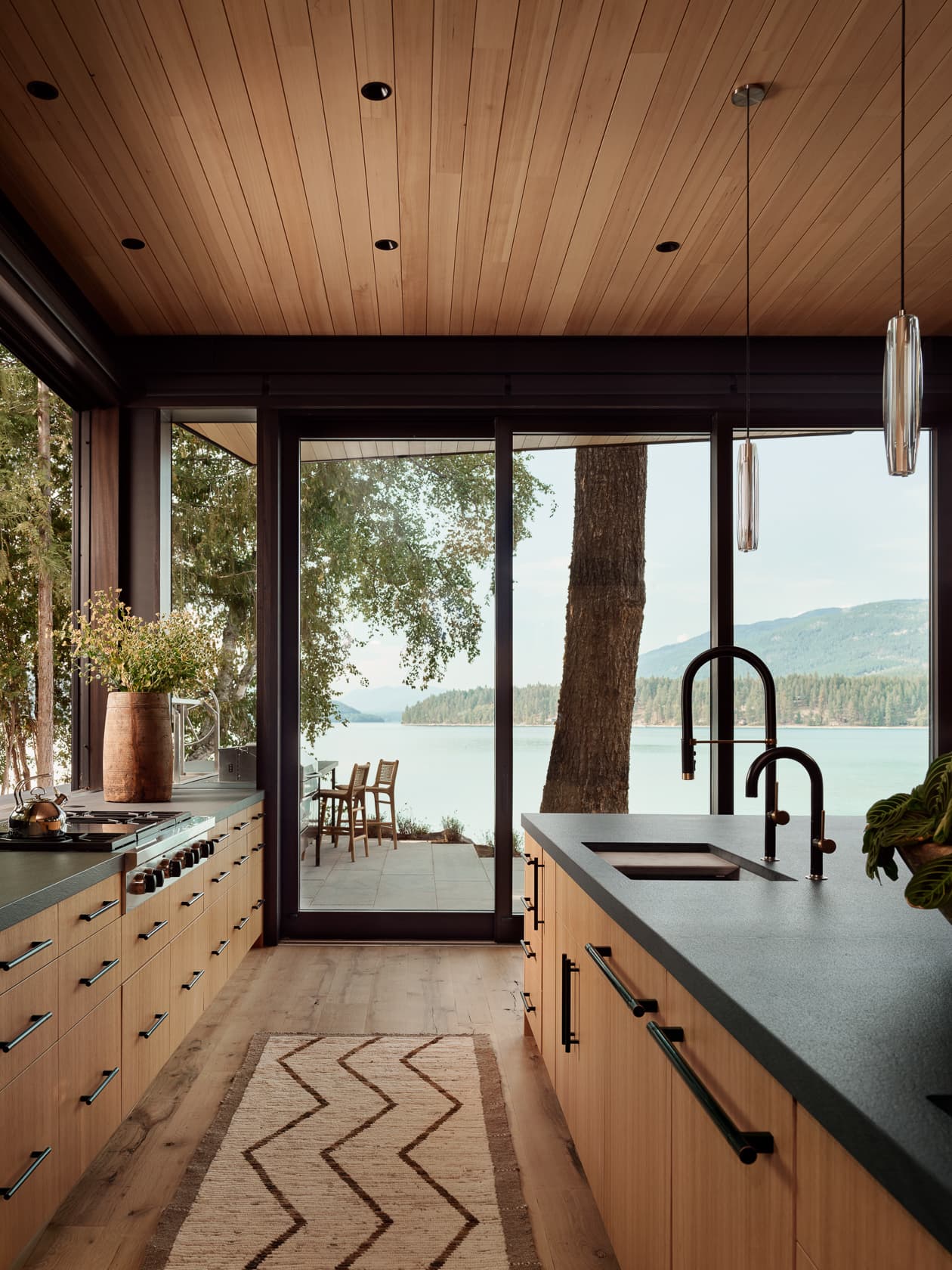 A modern kitchen with wood cabinets and a view of a lake through large glass doors.