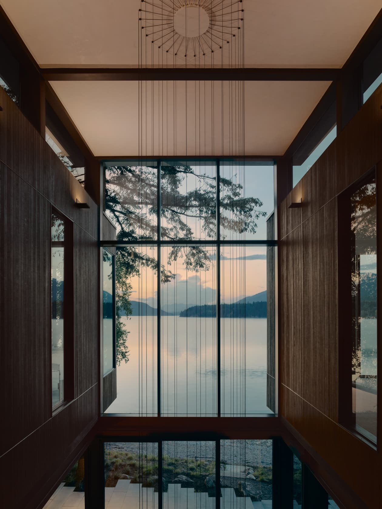 A tall, wood-paneled room frames a large window overlooking a calm lake and mountains at dusk.