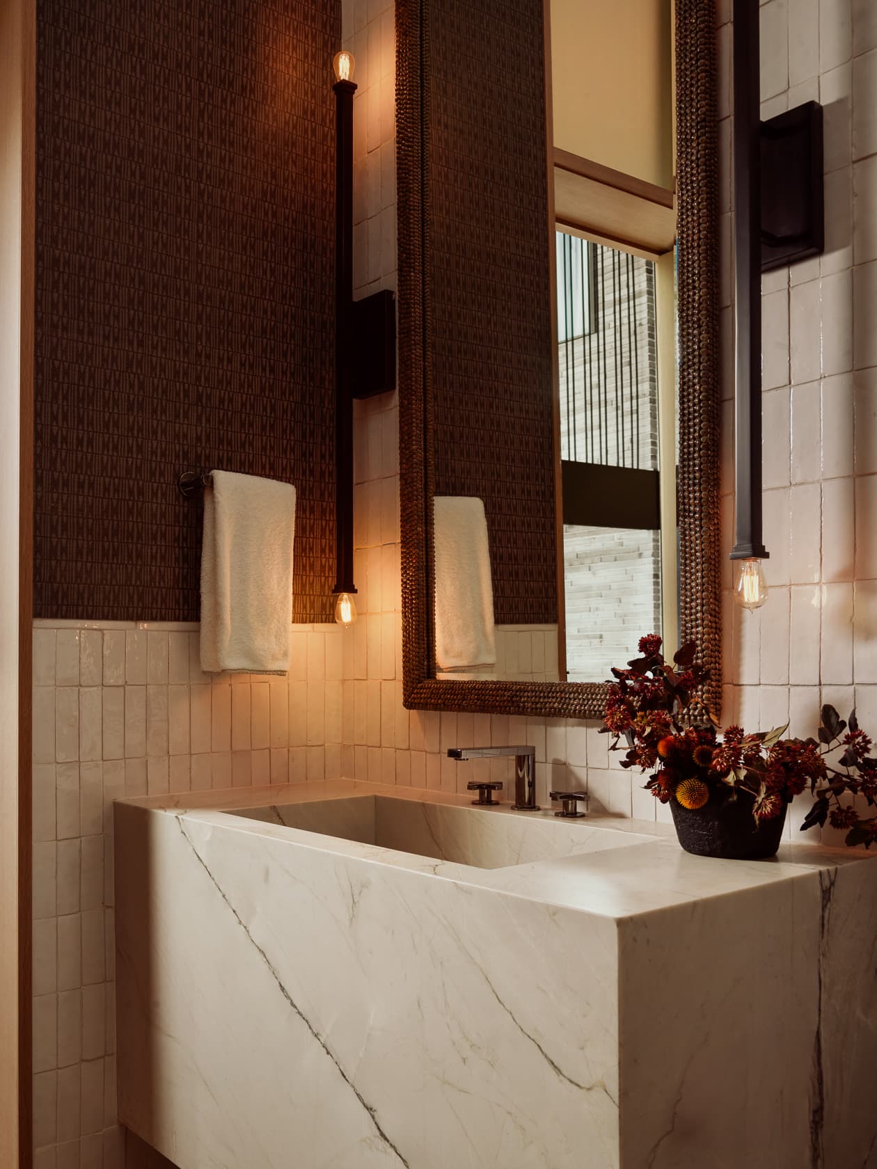 A modern bathroom with a marble block sink, dark patterned wallpaper, and warm vertical sconces.