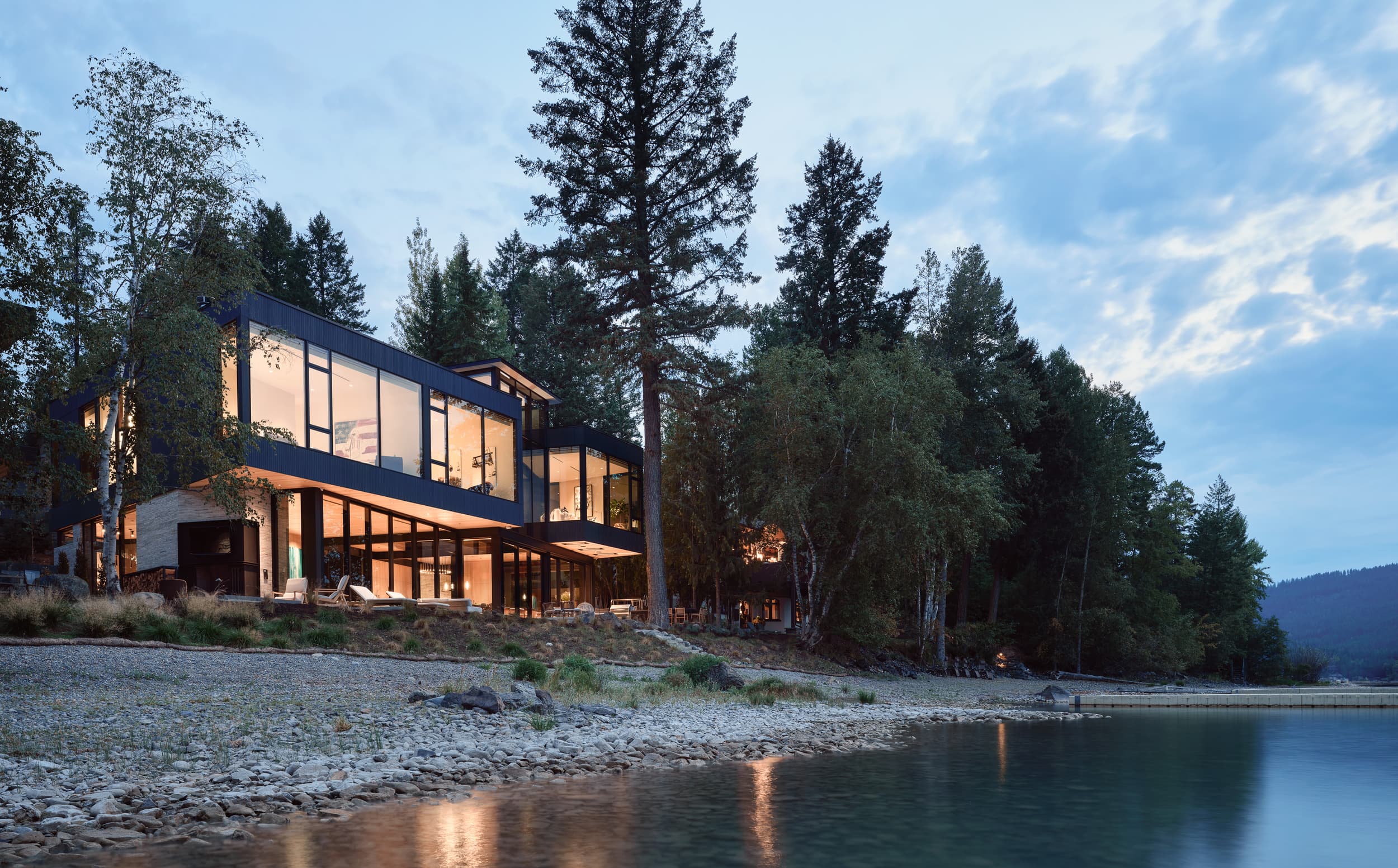 A modern, glass-walled house on a rocky lake shore at dusk, surrounded by a forest.