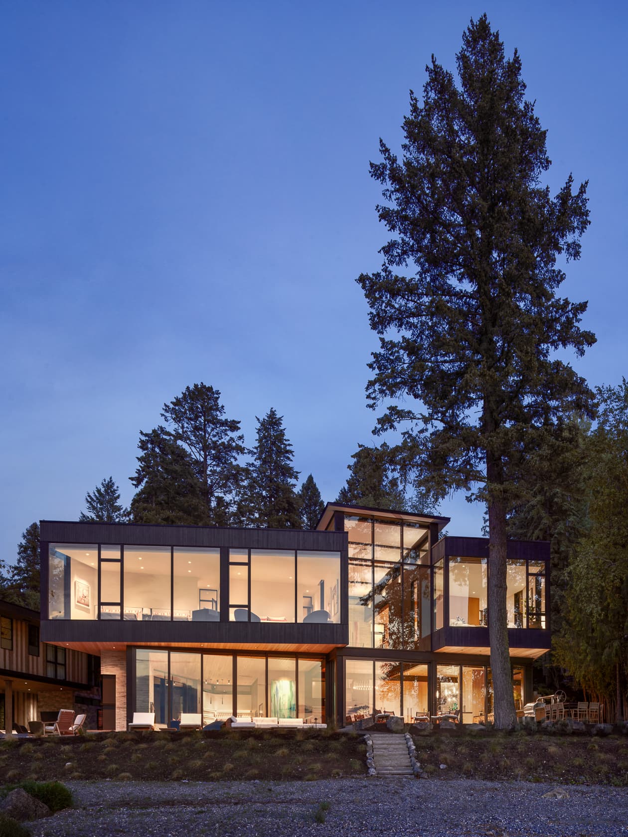 Photograph of a modern two-story home with glass walls, lit from within at dusk among tall trees.