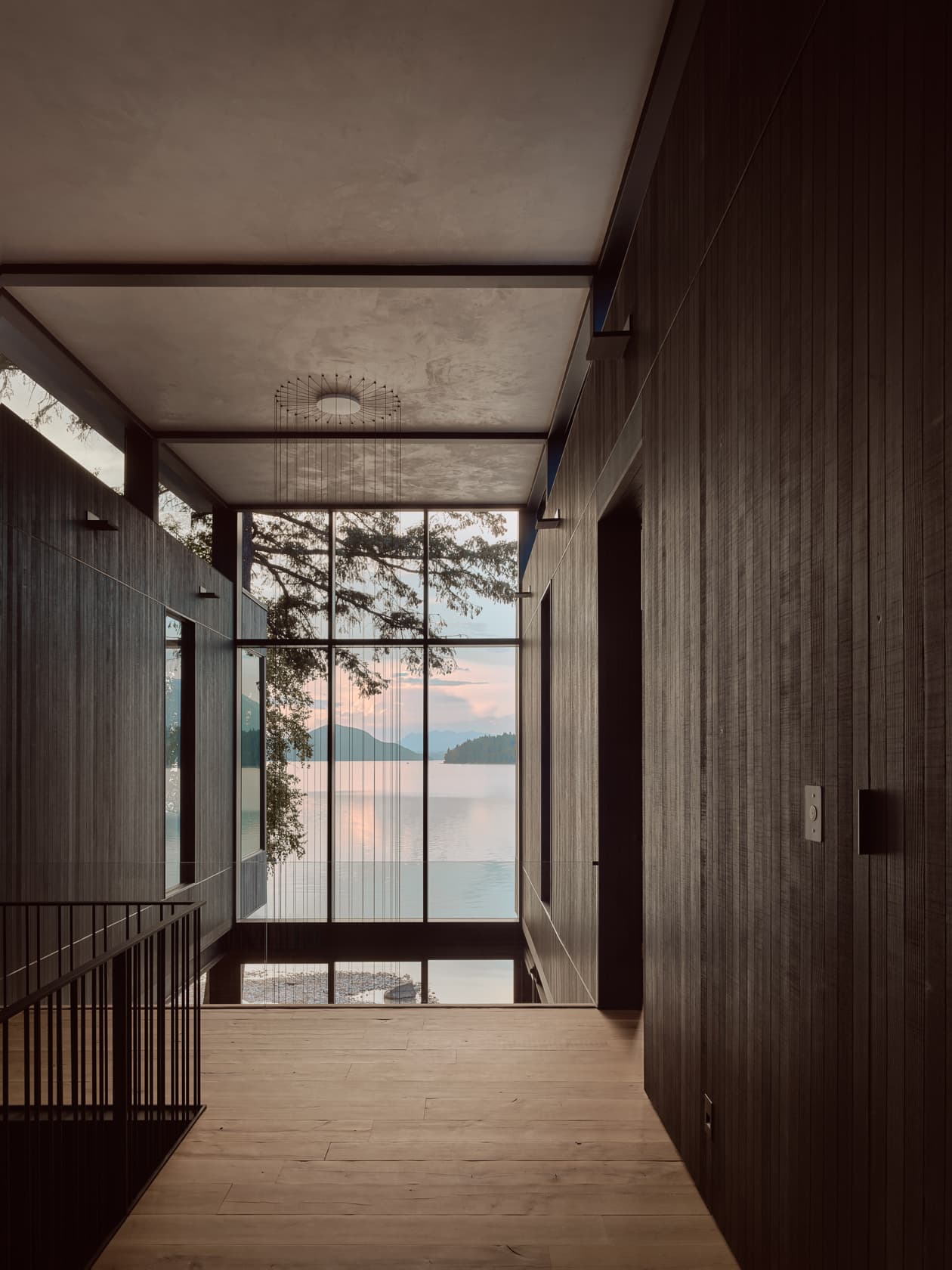 A modern, dark wood hallway leading to a large window overlooking a serene lake at sunset.
