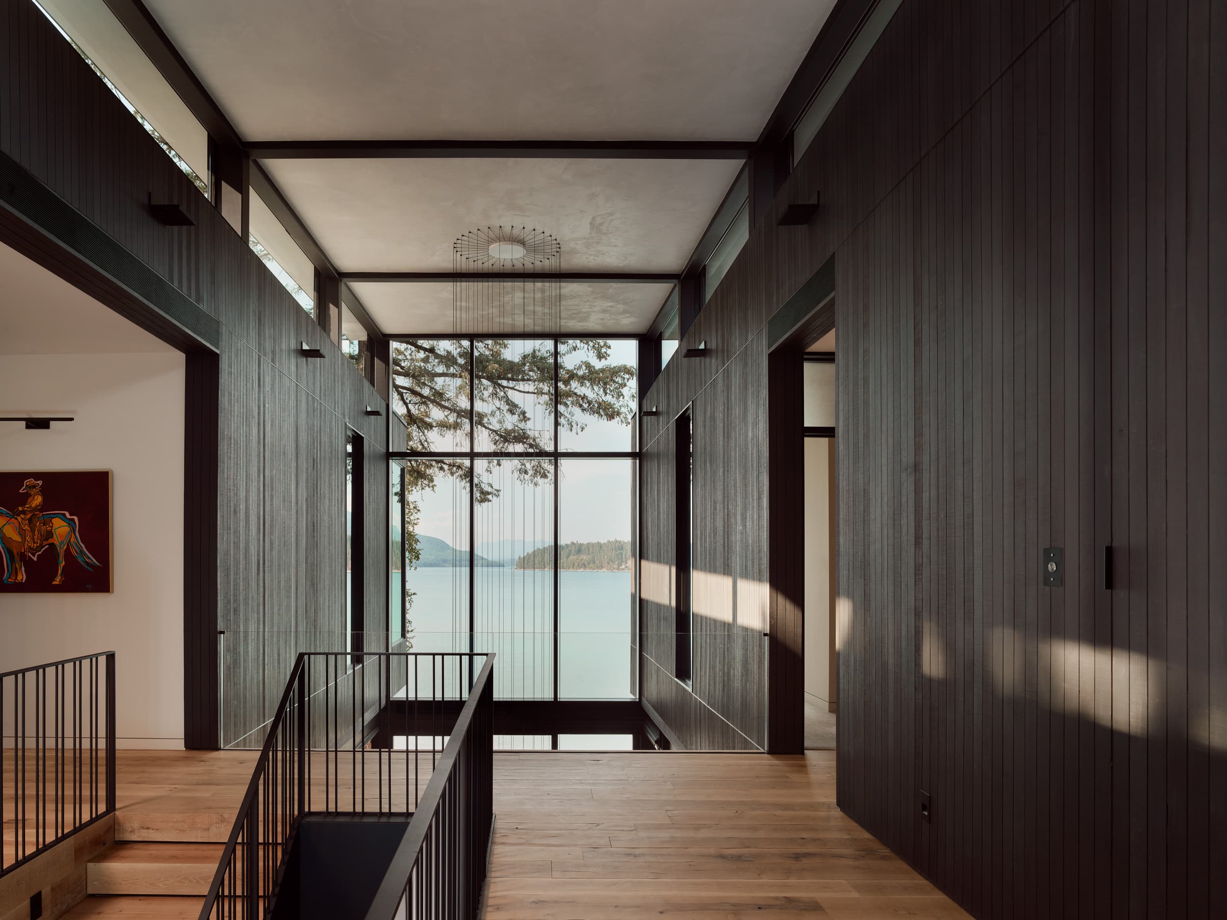 A dark wood-paneled hallway in a modern home overlooks a body of water through a large window.